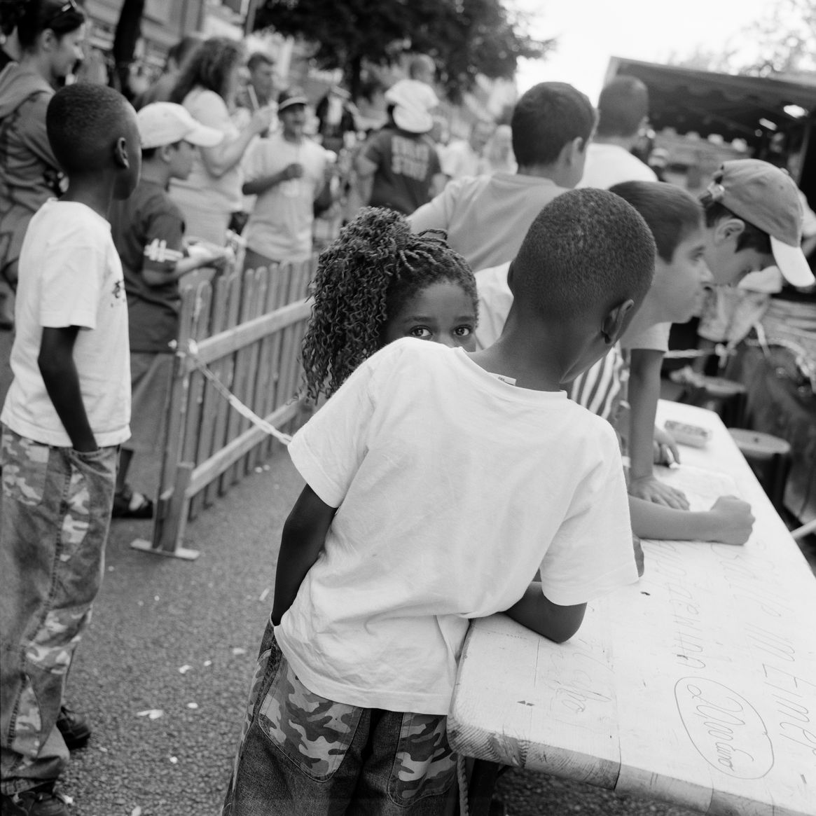 Black and white photograph: view of an outdoor party. Two children are standing next to a long table, one person is looking directly into the camera and is partially obscured by the person in front of them. 