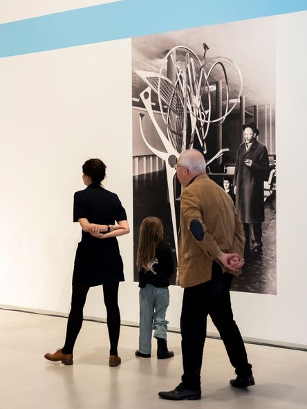 Foto: Drei Personen stehen vor einer schwarz-weiß-Fototapete, die Hans Uhlmann neben einer seiner Skulpturen zeigt.