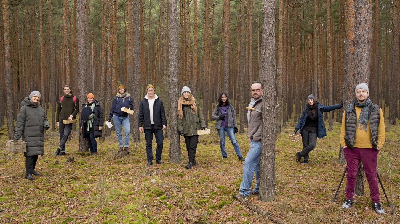 A group of people stand in a forest and look head-on into the camera.