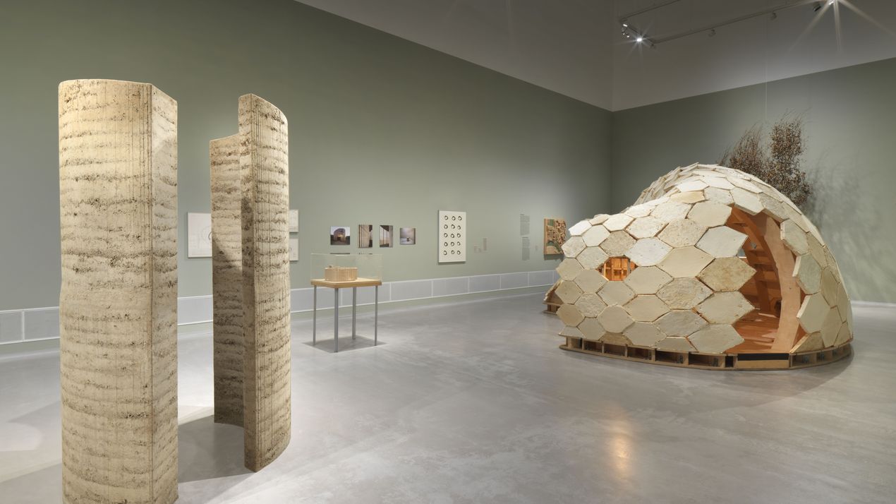 Exhibition view: View of a clay installation and a construction made of wooden mushroom modules. Further works and documentation of the participating projects hang on the matt green walls.