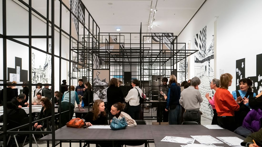 Foto: Blick in den Ausstellungsraum von „Kotti-Shop/SuperFuture" mit vielen Menschen, zwei langen Tischen und einer großen Gitterkonstruktion.