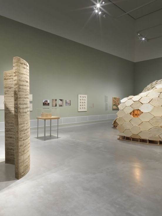 Ausstellungsansicht „Closer to Nature. Bauen mit Pilz, Baum, Lehm“, Berlinische Galerie Ausstellungsansicht: Blick auf eine Lehminstallation und eine Konstruktion aus Holz-Pilzmodulen. An den matt-grünen Wänden hängen weitere Arbeiten und Dokumentationen der beteiligten Projekte.