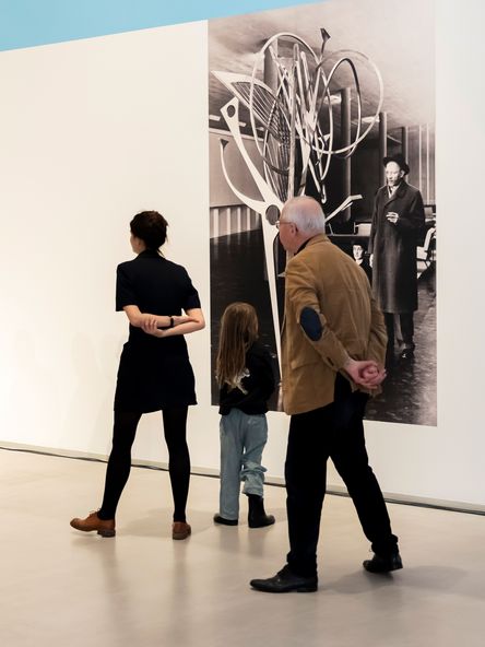 Photo: Three people stand in front of a black and white photographic wallpaper showing Hans Uhlmann next to one of his sculptures.