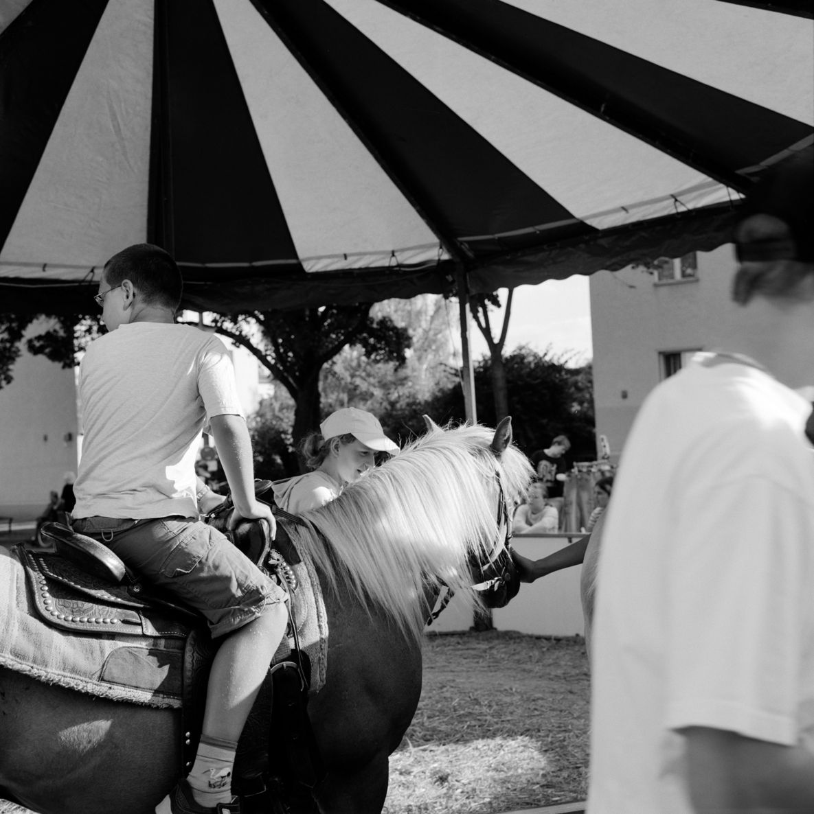 Black and white photograph: Side view of a boy on a horse under a striped canopy. Other people can be seen in the background.