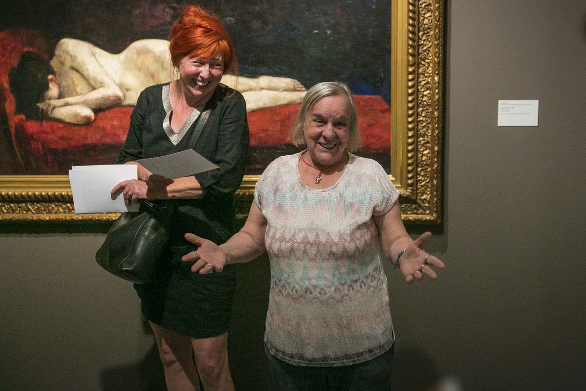 Foto: Eine größere Frau und eine kleinere Frau stehen vor einem Gemälde und blicken lachend in die Kamera. Die kleinere Frau hat ihre Arme einladend ausgebreitet.