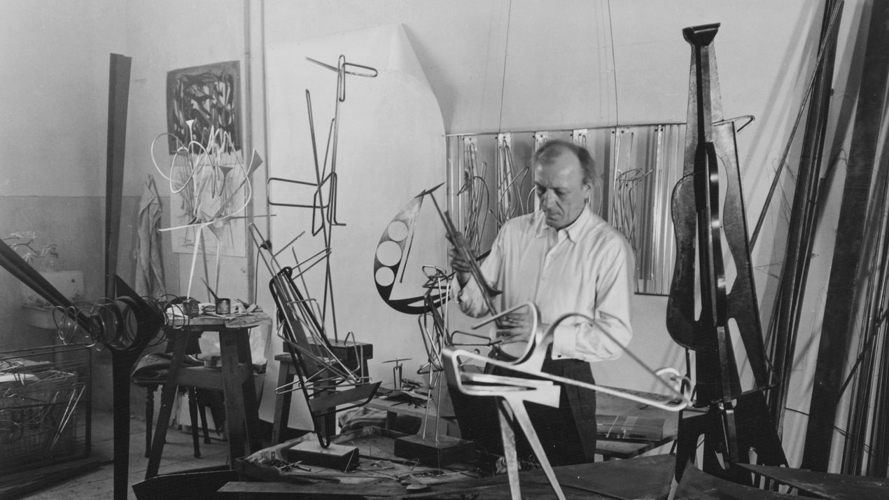 Black and white photograph of Hans Uhlmann in his studio. The artist is surrounded by his metal sculptures in progress.